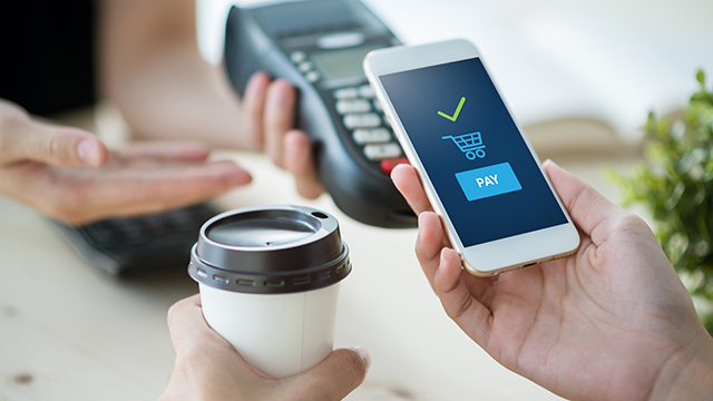 A person's hands holding a coffee and a phone with a screen that reads "pay" and another person holding a card reader.
